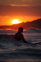 Silhouette of surfer riding wave during dramatic orange sunset, with water droplets splashing against dark sky and glowing sun setting behind mountains.
