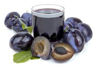 Fresh plum juice in glass surrounded by whole and cut ripe purple plums with green leaves, showcasing natural fruit beverage.