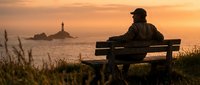 Solitary figure sitting on wooden bench overlooking coastal lighthouse at golden sunset, contemplative mood, peaceful seascape with warm sky tones.
