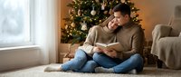 Young couple reading book together on floor near decorated Christmas tree in cozy living room during winter holidays celebration at home.