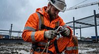 Professional construction worker in orange safety gear welding metal framework at industrial building site with protective equipment and hard hat.