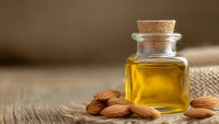 Golden almond oil in glass bottle with cork stopper and scattered almonds on rustic wooden surface for natural skincare and aromatherapy.