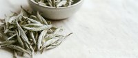 Dried white tea leaves scattered on linen surface with ceramic bowl, suitable for beverage packaging, wellness branding, and herbal product marketing.
