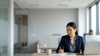 Woman in business attire writing at desk in modern office environment, suitable for corporate, professional, and workplace-related projects.