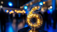 Glowing number six illuminated with warm fairy lights against dark background with blue bokeh lights, creating festive celebratory atmosphere.