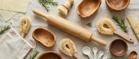 Wooden kitchen utensils and baking tools arranged on linen fabric with rosemary herbs, featuring rolling pin, spoons, bowls and pastry brushes.