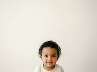 Toddler with curly dark hair and medium skin tone wearing white shirt, smiling at camera against neutral background. Suitable for parenting, childcare, family and education content.