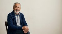 Senior man with gray hair and beard, Caucasian ethnicity, wearing navy blazer and light blue shirt, smiling at camera while seated. Suitable for business, corporate, lifestyle and professional portraits.