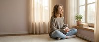 Young woman sitting cross-legged on floor by window holding coffee mug in cozy home interior with natural light and plants for relaxation lifestyle content.