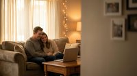 Young couple relaxing together on couch with laptop in cozy living room with warm lighting and string lights creating intimate atmosphere at home.