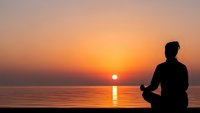 Man silhouette meditating in lotus pose at sunset by calm ocean water with golden sky reflection creating peaceful mindfulness scene for wellness content.