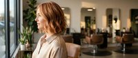 Professional woman with wavy brown hair in modern salon interior with styling chairs and plants, business portrait for beauty industry marketing.