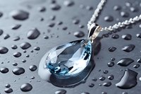 Elegant teardrop aquamarine crystal pendant necklace on silver chain against dark wet surface with water drops, macro jewelry photography with soft focus.