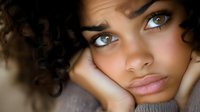 Young woman with curly dark hair and light eyes resting chin on hands, expressing sadness or contemplation. Use for mental health, wellness, emotional support, counseling services, or lifestyle content.