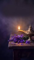 Ornate brass oil lamp with glowing flame beside purple lavender flowers on rustic wooden table against dark moody background for aromatherapy and relaxation.