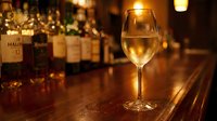 Glass of white wine on wooden bar counter in upscale restaurant with bottles in background creating warm ambient lighting atmosphere for dining experience.