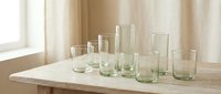 Collection of clear green-tinted drinking glasses in various sizes arranged on wooden table near window with natural light for kitchen and dining decor.