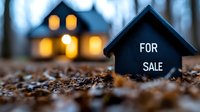 Small for sale sign in front of glowing house model at dusk, real estate concept with warm light and bokeh effect on autumn ground with fallen leaves.