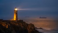 Lighthouse beacon illuminating twilight seascape with cargo ship on horizon. Coastal navigation safety tower on rocky cliff during golden hour sunset.