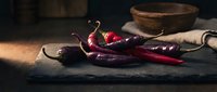 Fresh red and purple chili peppers on dark slate board with rustic wooden bowl, dramatic moody lighting for food and culinary concepts.