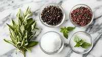Fresh herbs and seasoning ingredients on marble background with black peppercorns, pink peppercorns, sea salt and basil leaves arranged in glass bowls.