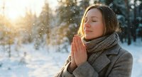 Young woman with closed eyes in prayer pose enjoying peaceful winter morning with falling snow and warm sunlight in snowy forest landscape.