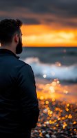 Bearded man in dark jacket gazing at dramatic ocean sunset with golden bokeh reflections on waves, creating atmospheric mood for contemplation and solitude.