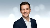 Professional young Caucasian businessman in navy suit smiling confidently against cloudy sky background for corporate marketing materials.