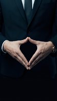 Professional businessman in dark suit making heart shape with hands showing care and compassion in corporate environment for business concepts.