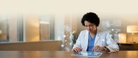 Young African American female doctor in white coat working on digital tablet with holographic medical icons and data visualization in modern hospital office.