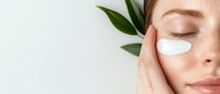 Young woman with natural makeup applying white moisturizing cream patches under eyes, green leaves decoration on white background, skincare concept.
