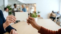 Two professionals exchanging blank business card in modern office environment with colleagues working in background, suitable for corporate communication and networking concepts.