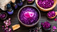 Purple bath salt with rose petals in wooden bowl, essential oils and dried flowers for aromatherapy spa treatment on dark wooden background. Natural wellness setup.