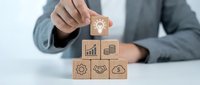 Professional businessman in suit arranging wooden blocks with business icons including lightbulb, charts, handshake, and gears on white desk for strategy planning.