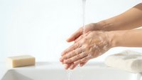 Hands washing under running water with soap bar nearby on white towel background for hygiene and cleanliness routine in bathroom or kitchen setting.