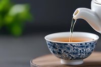 Pouring hot tea into traditional blue and white porcelain cup with floral pattern on wooden coaster against dark background with green bokeh effect.