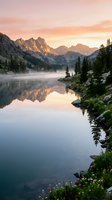 Serene alpine lake at sunrise with misty mountains, evergreen forest, and mirror-like water reflection. Ideal for nature, travel, and landscape design projects.