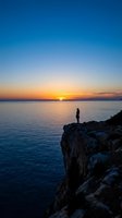 Woman standing on rocky cliff overlooking calm ocean at sunset, golden sky reflecting on water, serene coastal landscape for travel, wellness, and inspirational projects.