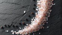 Pink Himalayan salt crystals scattered on dark slate stone background with black peppercorns creating diagonal composition for culinary and food styling.