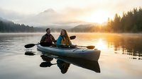 Couple kayaking on calm lake at sunrise surrounded by misty mountains and evergreen forest, adventure travel and outdoor recreation activity.