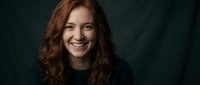 Happy young Caucasian woman with curly red hair smiling warmly at camera against dark background for portrait photography and lifestyle content.