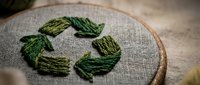 Hand-embroidered green recycling symbol on natural linen fabric in wooden hoop, showcasing sustainable eco-friendly craft and environmental awareness concept.