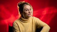 Young Caucasian woman with glasses and messy bun hairstyle wearing yellow turtleneck sweater smiling confidently against dramatic red background lighting.