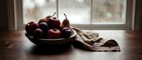 Rustic bowl of red apples and pears on a wooden table by a rainy window, moody still life with soft natural light and linen cloth.