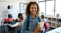 Young African American female teacher smiling in modern classroom holding books with diverse students studying at desks in bright educational environment.