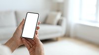 Two hands holding smartphone with blank white screen in modern living room interior with sofa and window light for app mockup.