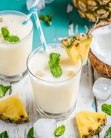 Fresh pina colada cocktails with coconut milk, pineapple pieces and mint leaves garnish on white wooden table. Tropical summer drink served with ice cubes.
