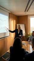 Professional blonde businesswoman presenting financial data charts to corporate team in modern conference room with wooden panels and natural lighting.