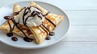 Homemade crepes with whipped cream, drizzled chocolate and caramel sauce served on white plate against rustic wooden background. Perfect for dessert menu.