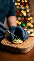 Chef hands in black gloves using culinary torch to caramelize appetizer on wooden board with festive Christmas lights bokeh background for holiday cooking.
